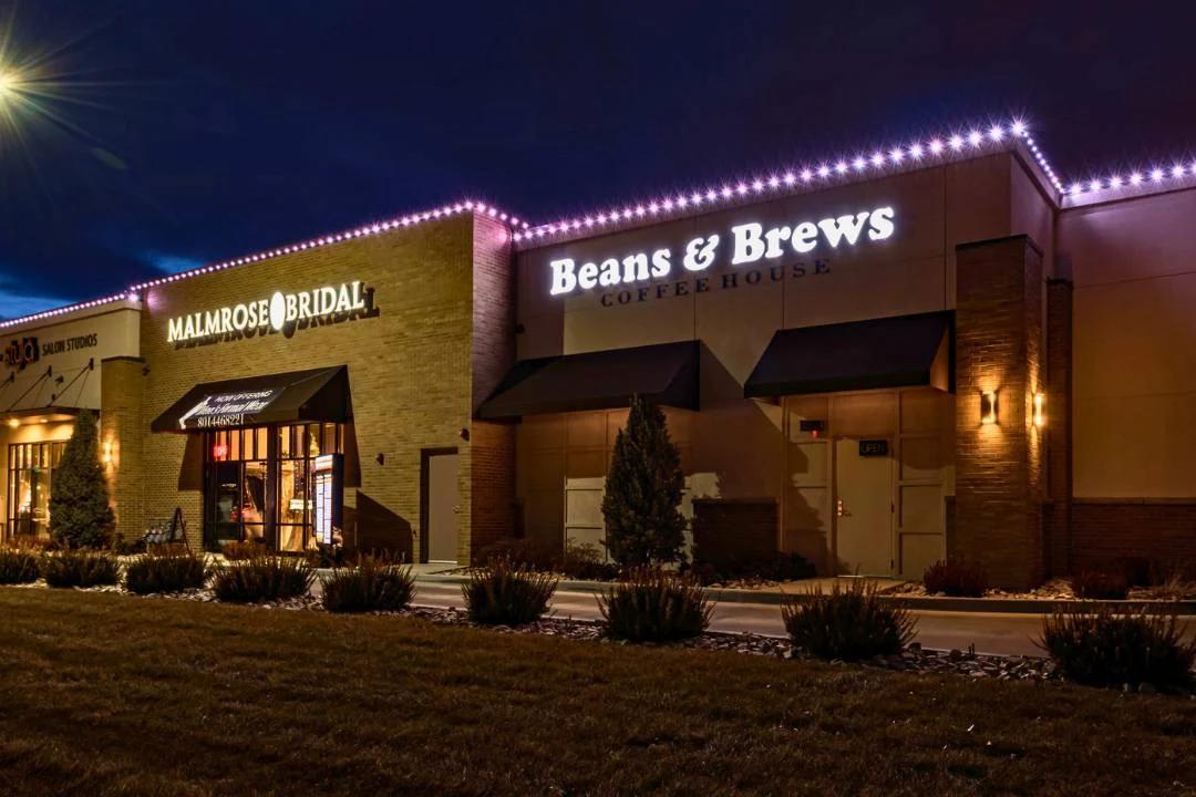 A brick building at night features illuminated signs for “Malmrose Bridal” and “Beans & Brews Coffee House,” with string lights along the roof and small bushes in front. {{brizy_dc_image_alt imageSrc=