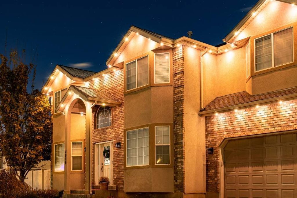A two-story house with brick and stucco exterior is lit by warm, white lights outlining the roof and windows at night. A tree with autumn leaves stands to the left, and the sky is dark and starry. {{brizy_dc_image_alt imageSrc=