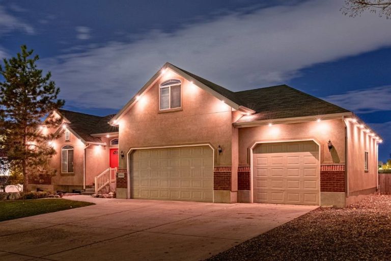 Single-story suburban house with beige walls, red brick accents, and a dark roof, illuminated by exterior lights at night. The home features a red front door, attached garages, and a concrete driveway. {{brizy_dc_image_alt imageSrc=