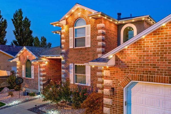Two-story red brick house with white trim, outdoor lighting along the roofline, arched windows, landscaped yard with shrubs and rocks, and a white garage door, pictured at dusk with a clear blue sky. {{brizy_dc_image_alt imageSrc=