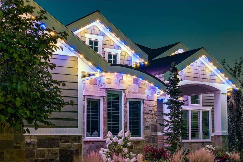 A modern house at dusk with stone and siding exterior, large windows, a front porch, and colorful string lights outlining the roof. Landscaping includes trees, shrubs, and white flowers in the garden. {{brizy_dc_image_alt imageSrc=