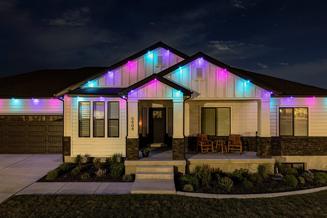 A modern single-story house at night, with colorful string lights in blue, purple, and pink decorating the roofline. The front porch has two wooden chairs, and the garden is neatly landscaped. {{brizy_dc_image_alt imageSrc=
