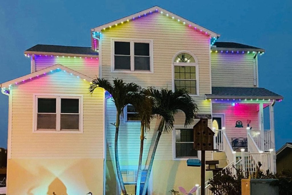 A two-story house decorated with multicolored string lights along the roof at dusk, with palm trees and a birdhouse in the front yard. {{brizy_dc_image_alt imageSrc=
