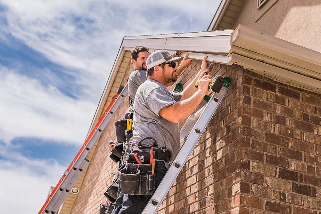 Two workers on ladders install or repair gutters on the roof edge of a brick house. They wear tool belts and gray shirts, working under a partly cloudy sky. {{brizy_dc_image_alt imageSrc=