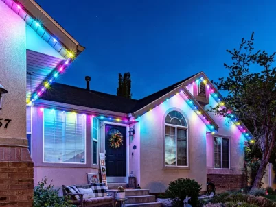 Attached image file A house at dusk decorated with colorful string lights along the roofline. The lights glow in blue, pink, yellow, and green. A wreath hangs on the front door, and a bench sits on the porch.