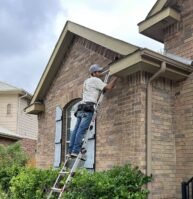 A person wearing a cap and tool belt stands on a ladder, working on the soffit of a brick house next to a bush, with another ladder positioned nearby under a cloudy sky.