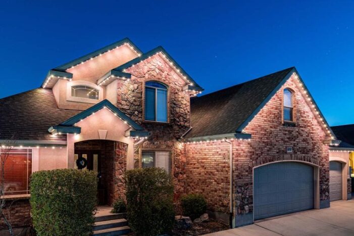 Two-story house with brick and stone exterior, lit by white string lights along the roofline, featuring a double garage, multiple windows, and neatly trimmed bushes in the front, photographed at dusk. {{brizy_dc_image_alt imageSrc=