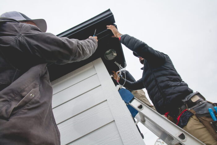 Two people stand on ladders installing or repairing something under the roof eaves of a white building, using tools and wires, with an overcast sky in the background. {{brizy_dc_image_alt imageSrc=