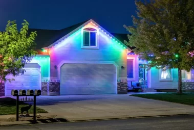 A suburban house at night is decorated with bright, colorful LED lights outlining the roof, illuminating the garage doors and front entrance. Trees, lawn, and mailboxes are visible in the foreground.