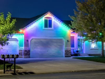 Attached image file A suburban house at night is decorated with bright, colorful LED lights outlining the roof, illuminating the garage doors and front entrance. Trees, lawn, and mailboxes are visible in the foreground.