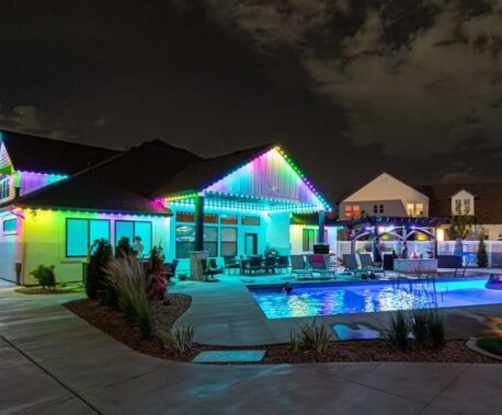 Attached image file A modern house at night with colorful LED lights outlining the roof, a lit swimming pool in the backyard, and outdoor seating surrounded by landscaping. The sky is partly cloudy.