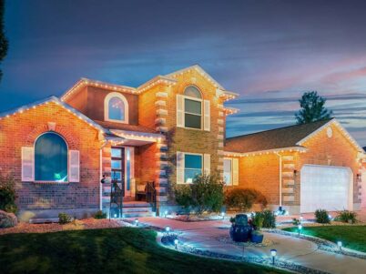Attached image file A modern brick house with large windows and decorative lights outlining the roof at dusk. The yard has landscaped plants, a curved walkway, and glowing pathway lights, creating a welcoming atmosphere.