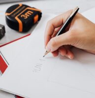 A hand sketching on white paper with a pencil next to a red folder, a tape measure, a laptop, and a black marker on a white desk.