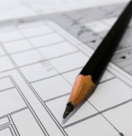 A sharpened black pencil rests on top of a detailed architectural blueprint with lines, grids, and measurements visible.